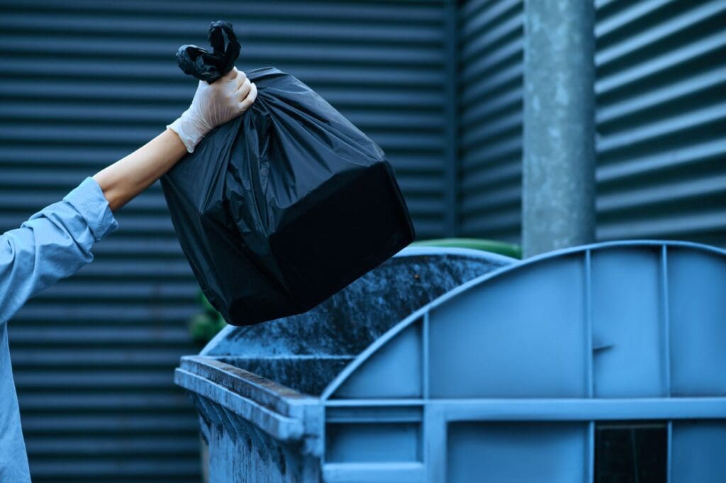 Persona tirando una bolsa negra de basura al contenedor de residuos. 
