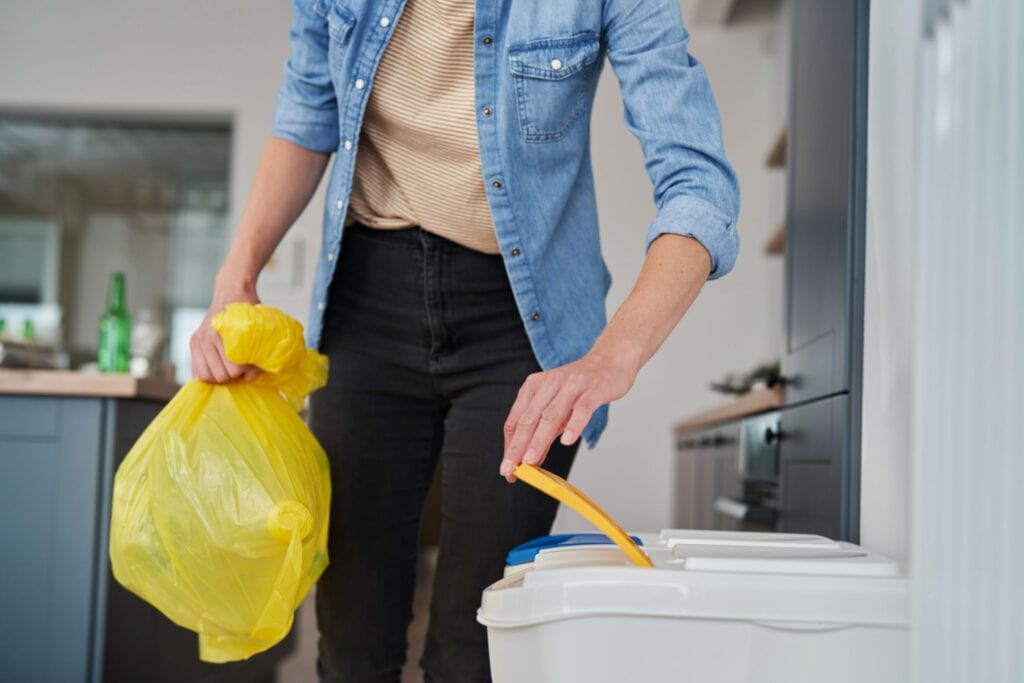 Persona tirando una bolsa de basura amarilla al contenedor de plásticos.