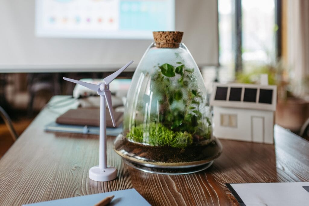 Modelo de turbina eólica y casa con paneles solares en una mesa, junto a un terrario de vidrio con plantas. La escena refleja conceptos de energía renovable y sostenibilidad en un ambiente de oficina.