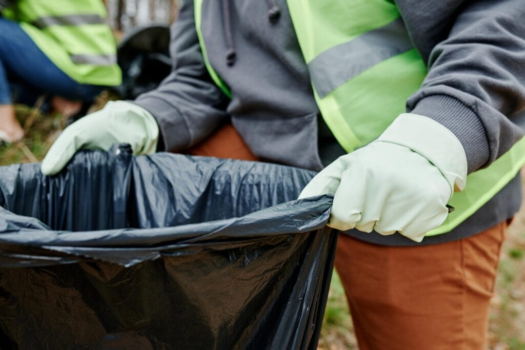 Persona con chaleco de seguridad y guantes sosteniendo una bolsa de basura negra durante una actividad de limpieza comunitaria al aire libre.