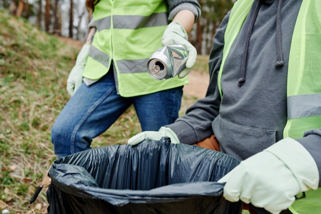 Dos personas con chalecos de seguridad y guantes, una de ellas sosteniendo una lata vacía y la otra una bolsa de basura negra, durante una actividad de limpieza comunitaria en un área natural.