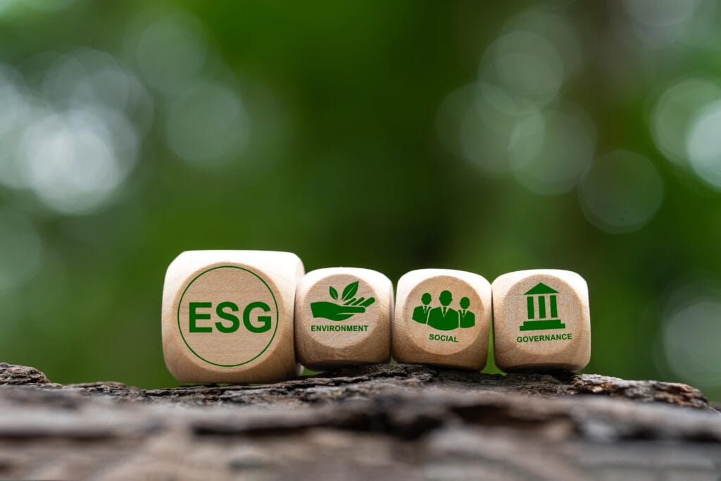 
La imagen muestra cuatro dados de madera colocados sobre una superficie natural, cada uno con símbolos y palabras relacionados con el concepto ESG (Environmental, Social, Governance). El primer dado tiene las letras "ESG" en color verde, mientras que los siguientes tres dados representan los componentes del ESG: el primero con una hoja para "Environment" (Medio Ambiente), el segundo con figuras humanas para "Social" (Social) y el tercero con un edificio que simboliza "Governance" (Gobernanza). El fondo es difuminado con tonos verdes, lo que sugiere un entorno natural y refuerza el enfoque en la sostenibilidad.