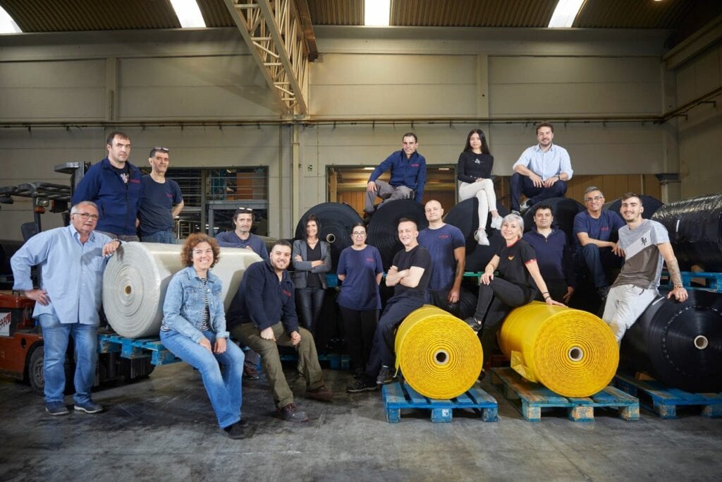 La imagen muestra al equipo de Plasticbag, fabricante de bolsas de basura recicladas y reciclables, posando juntos en el interior de una fábrica. El grupo está compuesto por alrededor de 15 personas, algunos de pie y otros sentados sobre grandes rollos de material industrial. Al fondo se observan más rollos y maquinaria relacionada con la fabricación. El ambiente es industrial y laboral, con iluminación propia de una nave de producción. El equipo viste ropa casual, algunos con uniformes de trabajo, reflejando un entorno colaborativo y profesional.