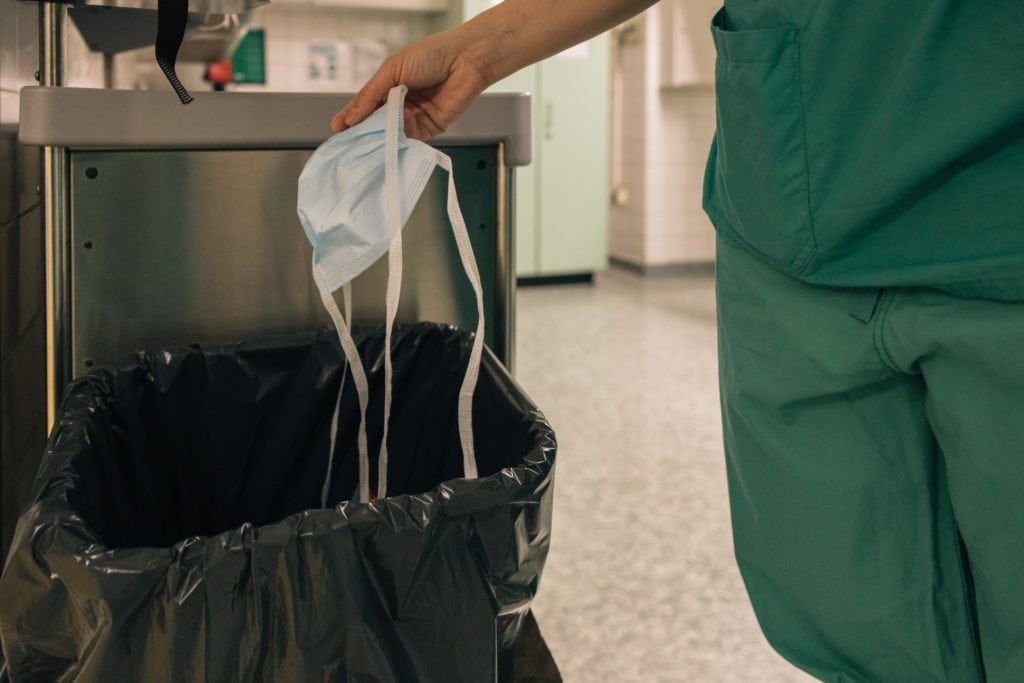 
La imagen muestra a un trabajador de la salud vestido con un uniforme verde arrojando una mascarilla desechable a un cubo de basura forrado con una bolsa de basura negra. El gesto refleja prácticas de eliminación segura de desechos médicos o potencialmente contaminados.