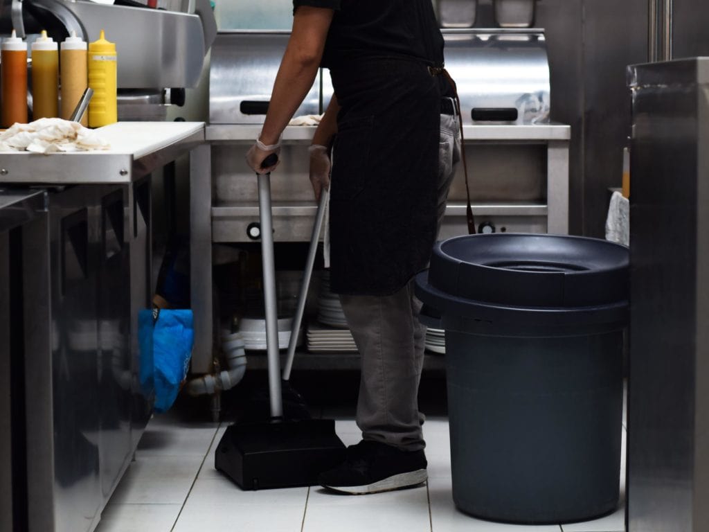 Empleado de restaurante limpiando el suelo cerca de una estación de trabajo con botellas de aceite, subrayando la necesidad de mantener la higiene en áreas de trabajo con residuos grasos.