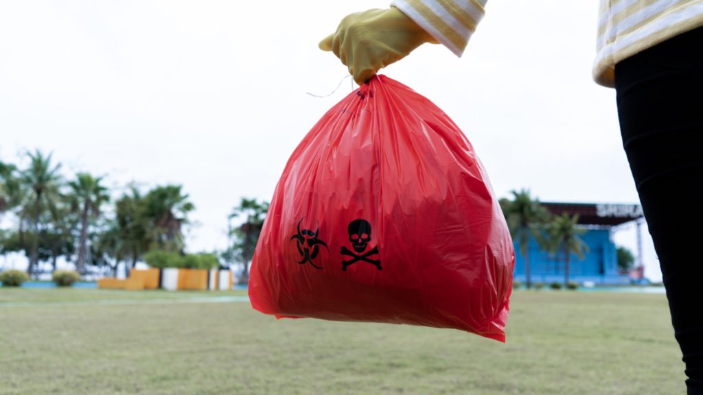 Persona sosteniendo una bolsa roja con desechos peligrosos, marcada con símbolos de advertencia, en un entorno al aire libre.