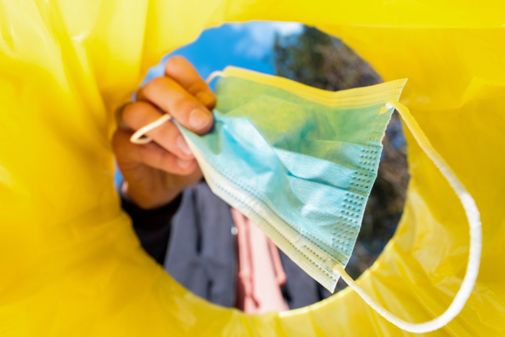 Persona tirando una mascarilla médica en un contenedor amarillo de residuos, resaltando la importancia de la correcta gestión de desechos sanitarios.