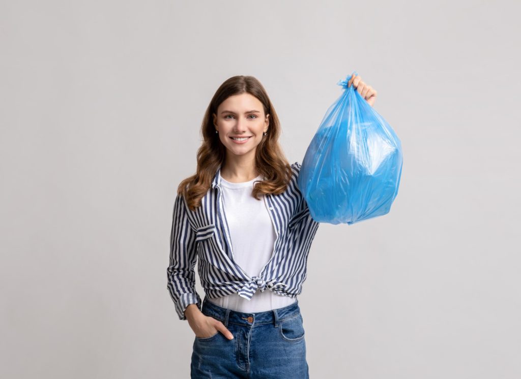 Mujer sonriente de pie sosteniendo una bolsa de plástico azul llena de residuos, sobre un fondo gris claro.