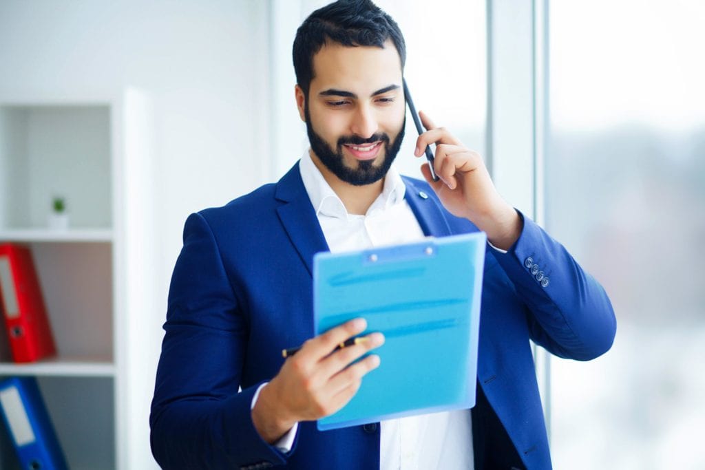 Hombre de negocios con traje azul hablando por teléfono mientras consulta un informe en un portapapeles, en una oficina luminosa.