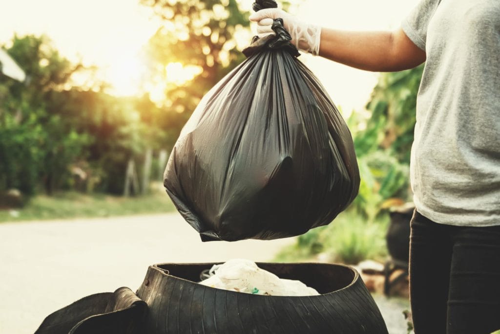 Mano con guante de limpieza profesional sosteniendo una bolsa de basura negra resistente junto a un contenedor urbano al atardecer.