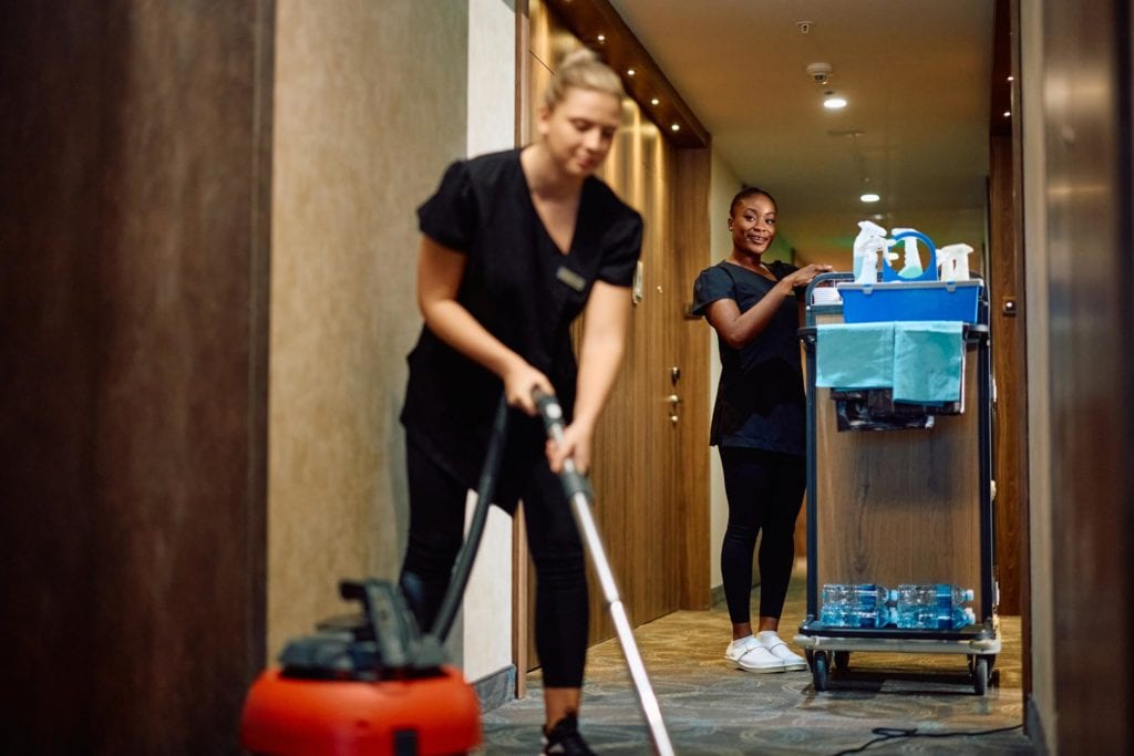 “Dos camareras de hotel con uniforme de limpieza, una empujando un carro de servicio y la otra aspirando el pasillo de una habitación.