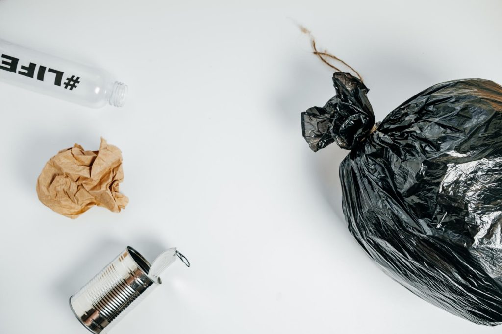 Bolsa negra de basura junto a botellas de plástico vacías, lata de metal y papel arrugado sobre fondo blanco, ilustrando la segregación de residuos