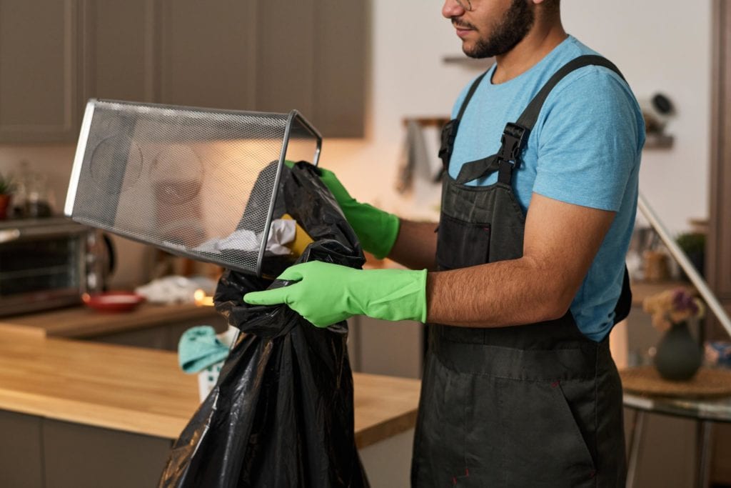 Hombre con guantes verdes vaciando una papelera de malla en una bolsa de basura negra en cocina, ilustrando uso intensivo de bolsas en verano.