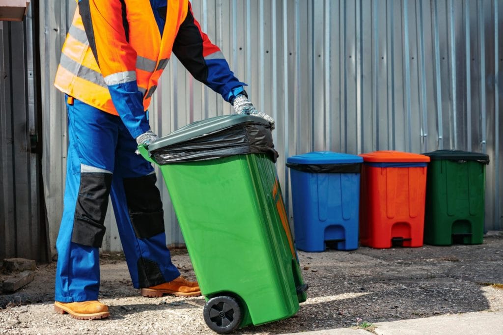 Trabajador con chaleco reflectante moviendo un contenedor de basura verde, con otros cubos de reciclaje de colores al fondo, junto a una pared metálica.