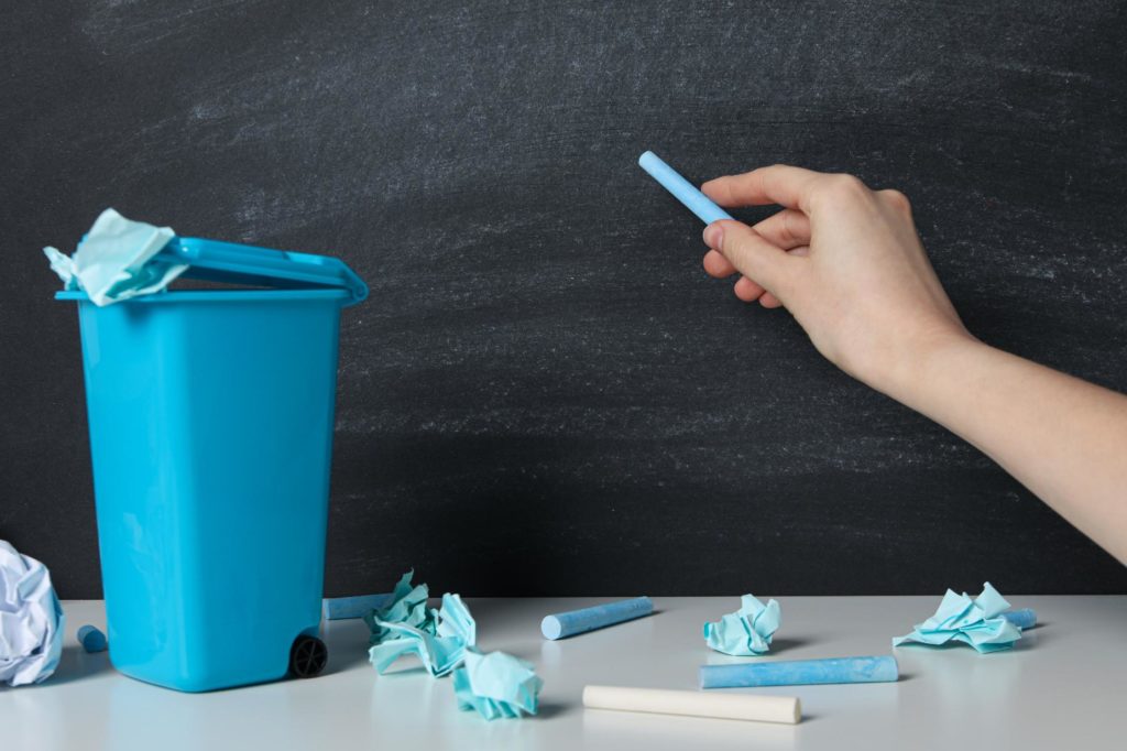 Papel arrugado y tizas junto a un contenedor azul para reciclaje en un aula escolar, fomentando la separación de residuos y hábitos sostenibles