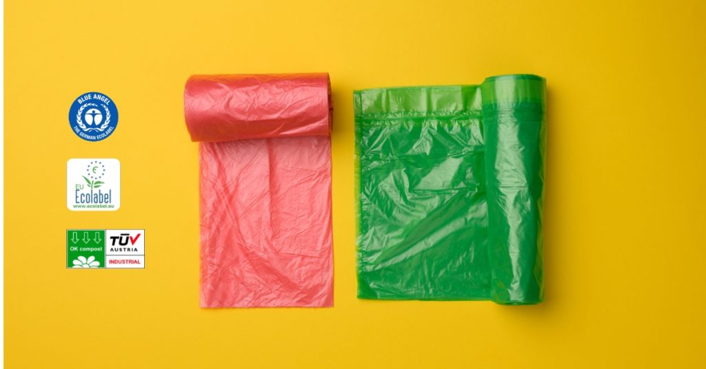 Rollos de bolsas de plástico recicladas y compostables en color rojo y verde, junto a los logotipos de las certificaciones Blue Angel, EU Ecolabel y OK Compost sobre fondo amarillo.