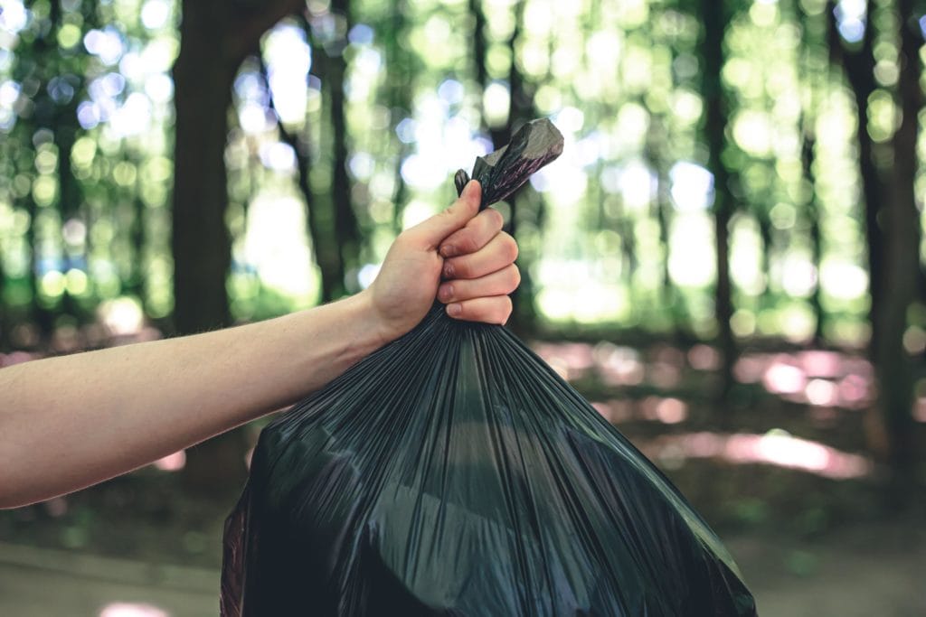 La mano de una persona sujeta con firmeza la parte superior de una bolsa de basura de plástico negra llena contra un fondo borroso de árboles verdes en un bosque o parque.